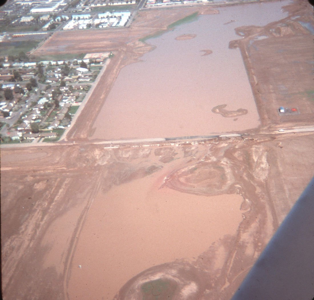 An aerial view showing a construction site with large muddy areas, some crisscrossing dirt roads, and a small residential neighborhood with houses and streets in the upper left corner.