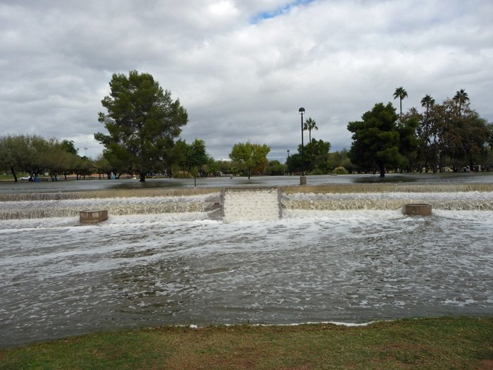 A flooded park with water flowing over a small dam, surrounded by trees and cloudy sky.