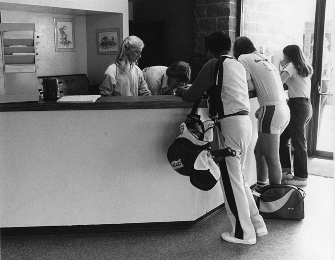 A group of people, mostly in athletic clothing, standing at a reception desk checking in or registering for an event, with some luggage and sports gear nearby.