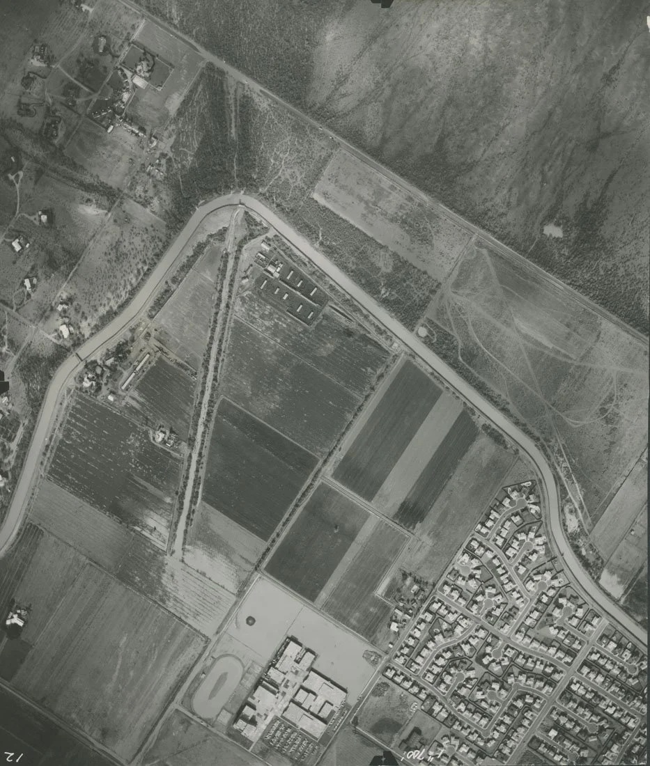 Aerial black and white photo of a rural area with farmland, roads, residential neighborhoods, and a school or public building complex.