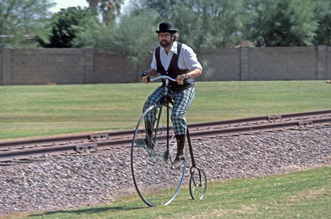 Man wearing vintage clothes riding a penny-farthing bicycle along a grassy area next to railroad tracks.