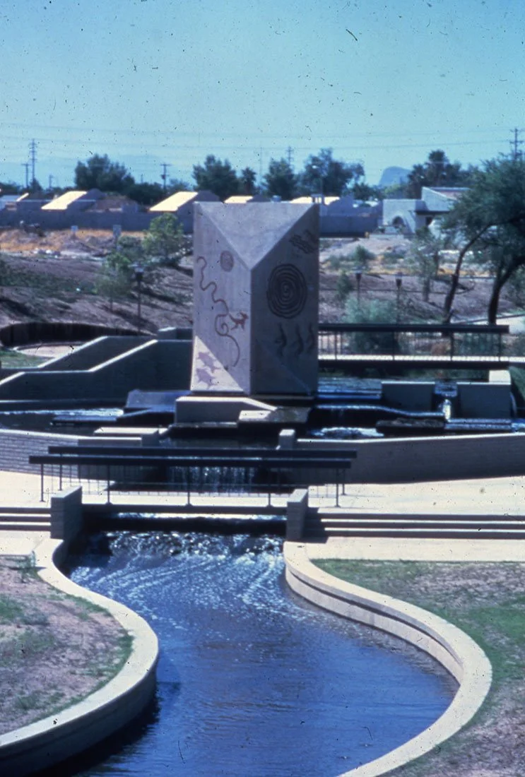 Public park featuring a water fountain with a curved stream of water, a decorative rectangular sculpture with abstract designs, and graffiti, with residential houses and trees in the background under a clear sky.
