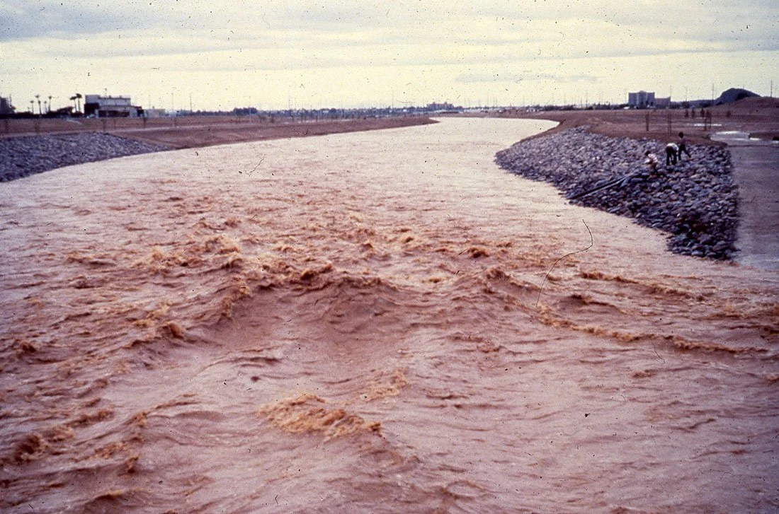 Flood waters flowing across a concrete channel in an urban area.