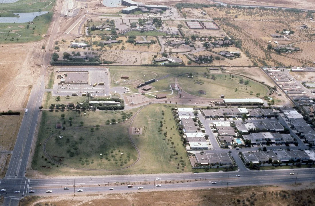 Aerial view of a suburban neighborhood featuring a park with grass, trees, and pathways, surrounded by residential and commercial buildings, roads, and open land.