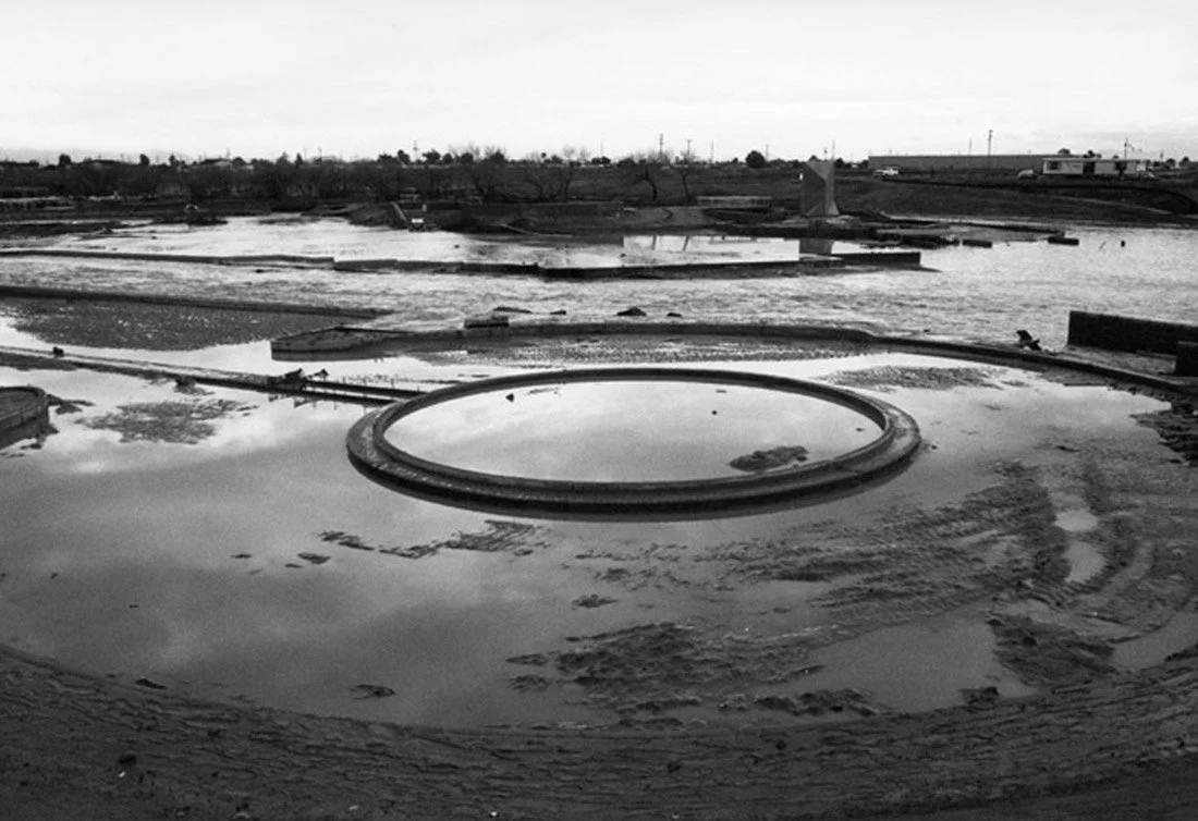 A circular pool surrounded by a muddy area, with multiple rectangular water features in the background, outdoors in a flat landscape.