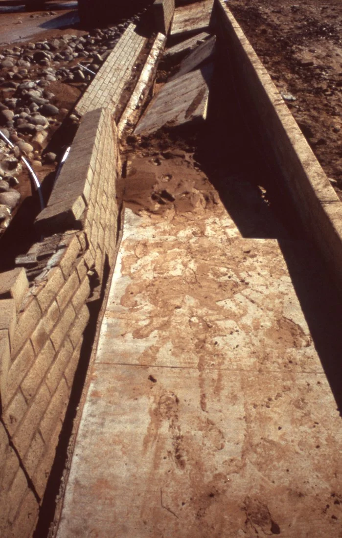 A partially constructed or damaged brick and concrete drainage trough or channel with uneven surfaces and debris, situated outdoors on a dirt surface.