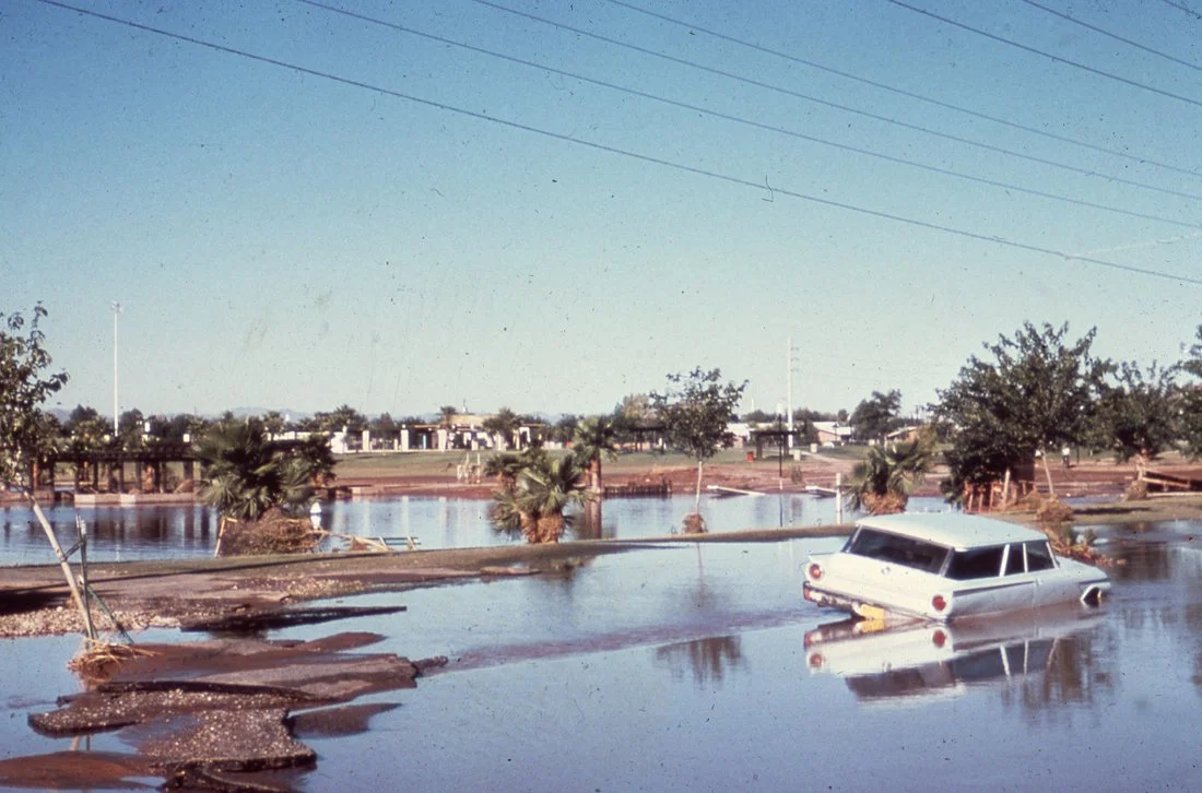 Flooded area with an submerged white car, trees, power lines, and a bridge over water.