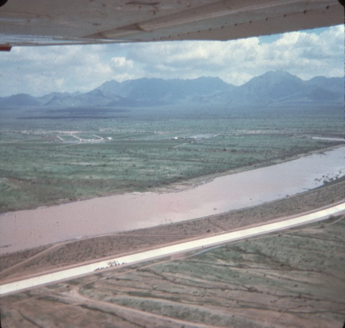 Aerial view of a landscape with mountains in the background, a river, and farmland with roads, seen from an airplane window.