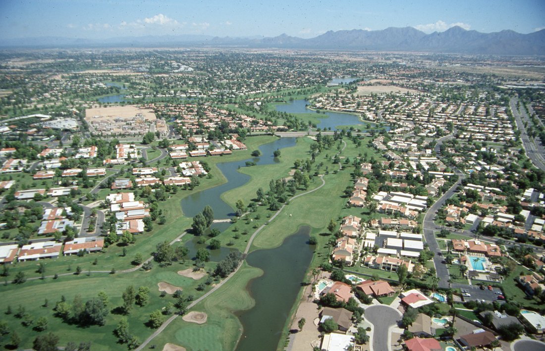 Aerial view of a residential area with a golf course, intersected by rivers, with mountains in the distance under a blue sky.
