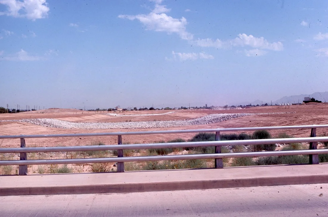 A barren, sandy landscape seen from a highway with a metal guardrail in the foreground, under a partly cloudy sky.