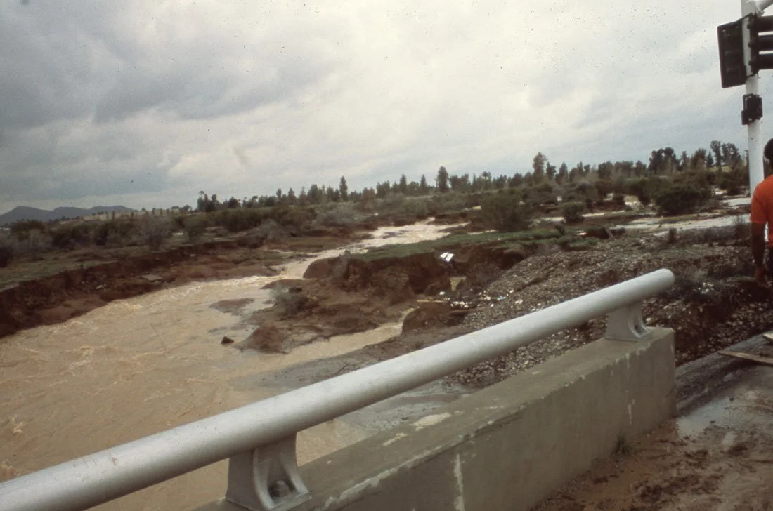 Damaged road with a broken bridge over a flooded, muddy river, and a cloudy sky in the background.