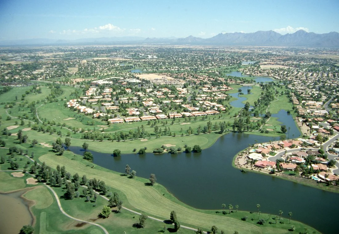 Aerial view of a suburban neighborhood with a golf course and water bodies, with a cityscape and mountains in the background.