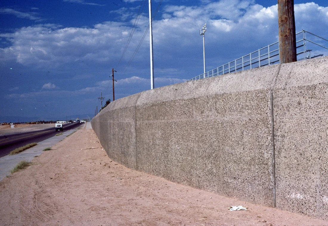 A concrete wall runs parallel to a road with a sidewalk, utility poles, and electric lines, under a partly cloudy blue sky.