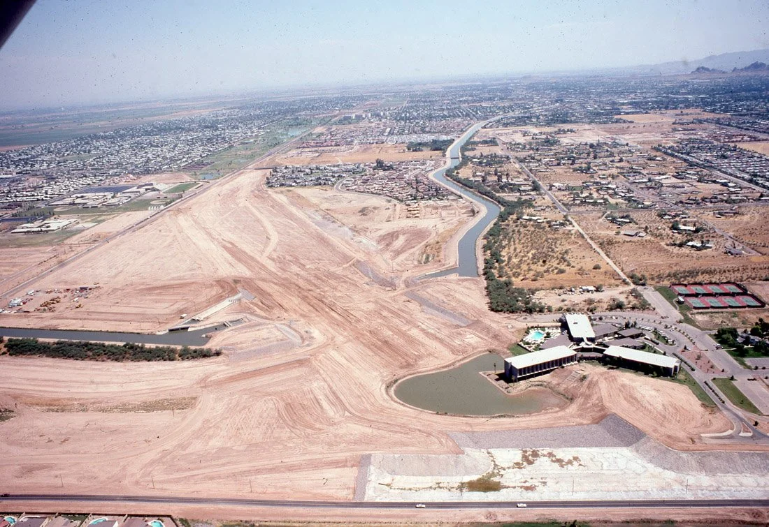 Aerial view of a construction site with dirt roads, a small water body, and a modern building, with a cityscape and mountains in the background.
