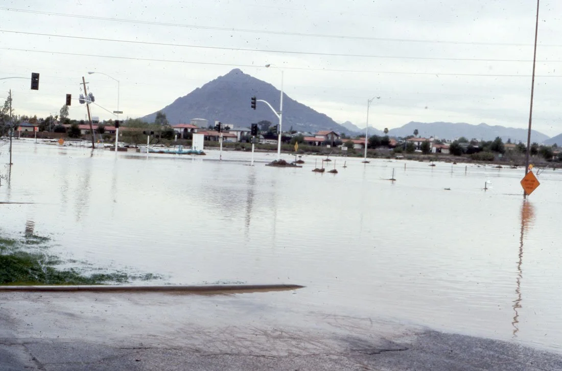 Flooded street with submerged traffic lights, signs, and power lines, mountain in the background, cloudy sky.