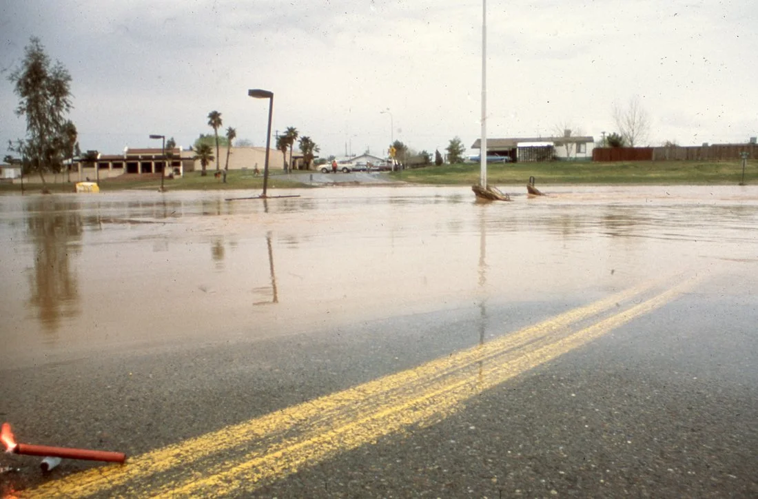 A flooded street with water covering the road, flooded lampposts, and submerged trees. Buildings and palm trees are visible in the background under a cloudy sky.