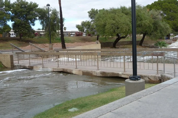 Flooded park area with a small bridge, trees, and lamp posts, water overflowing onto pedestrian walkway.