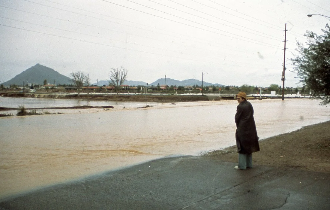 A person wearing a coat and sunglasses standing on a flooded street with water covering the road, with mountains and trees in the background.