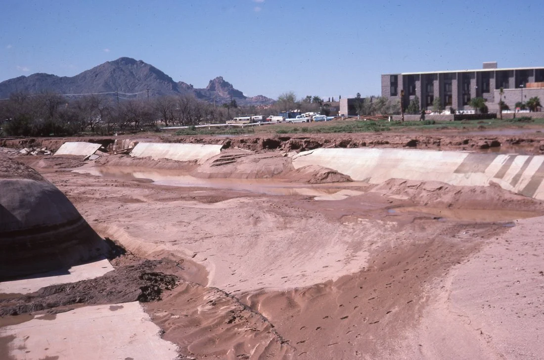 A partially dried and eroded flood control channel with concrete walls, with mountain range in the background and a modern building on the right. Several cars are parked in the distance.
