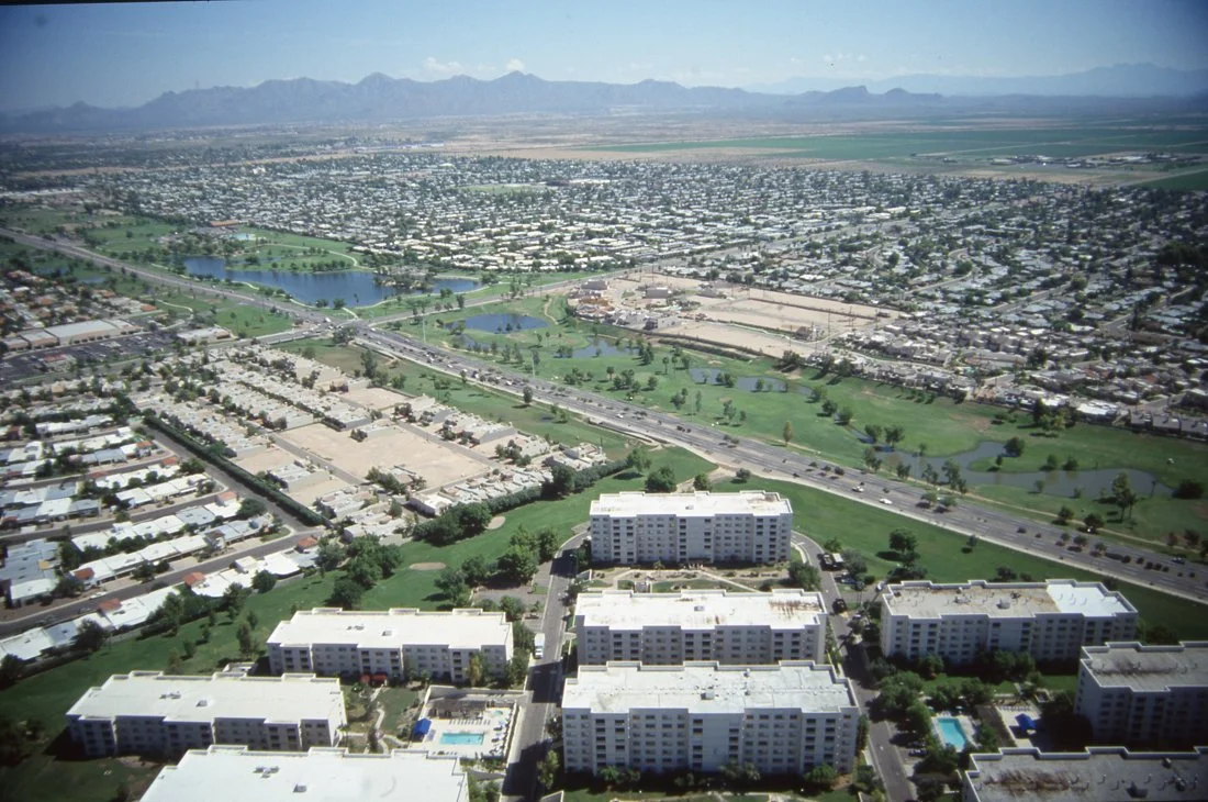 Aerial view of a city with residential buildings, parks, lakes, and a highway, with mountains in the distance.