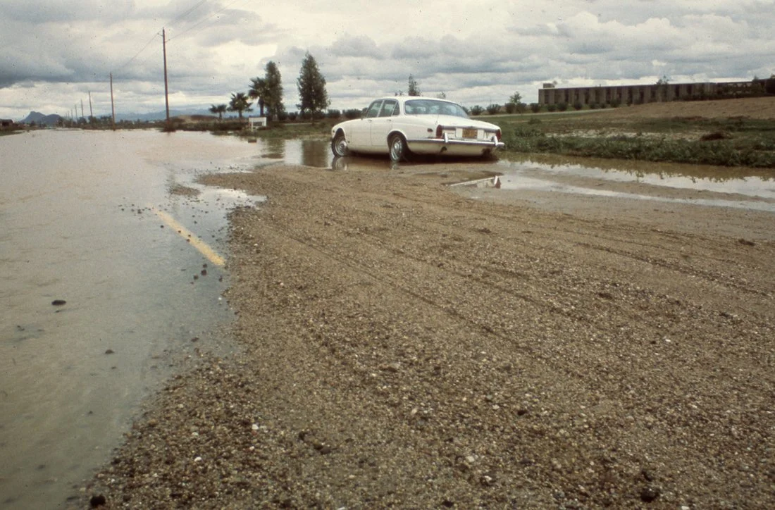 A white vintage car partially submerged in floodwaters on a gravel road with overcast sky and sparse vegetation.