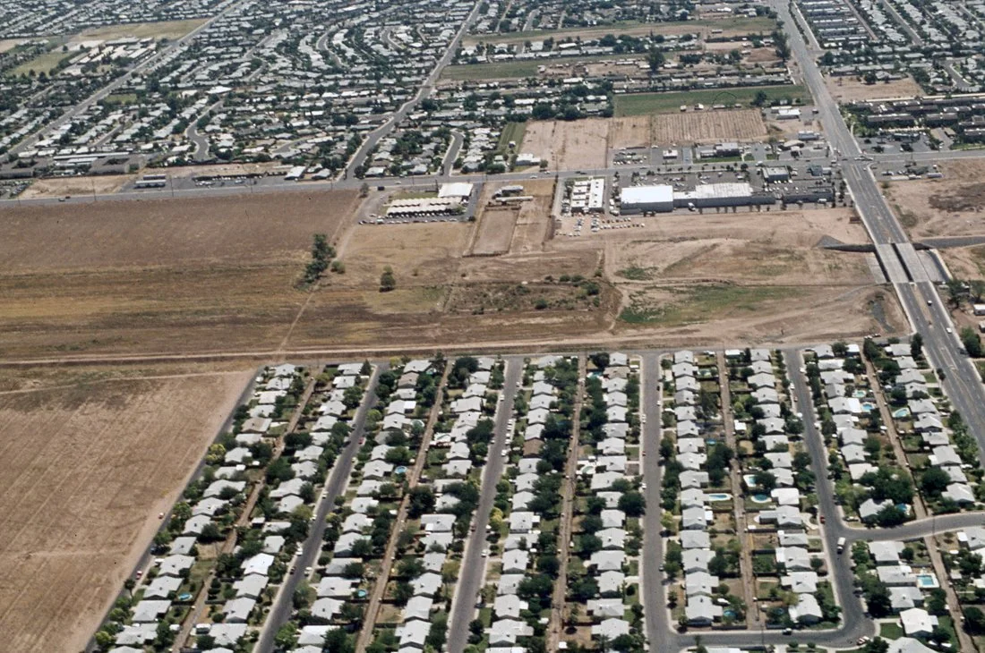 An aerial view of a suburban neighborhood with rows of houses, a commercial area, and open land with dirt roads and train tracks.