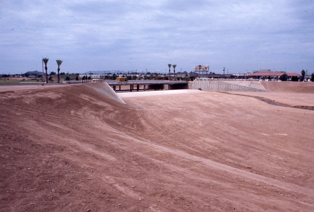 A dry, dusty canal or irrigation ditch with a concrete wall on one side and a bridge in the background. There are a few palm trees and some buildings visible in the distance under a mostly cloudy sky.