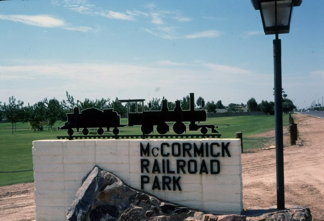 Sign for McCormick Railroad Park with a silhouette of a train on top, situated on a grassy area with trees, a dirt path, and a street lamp nearby.