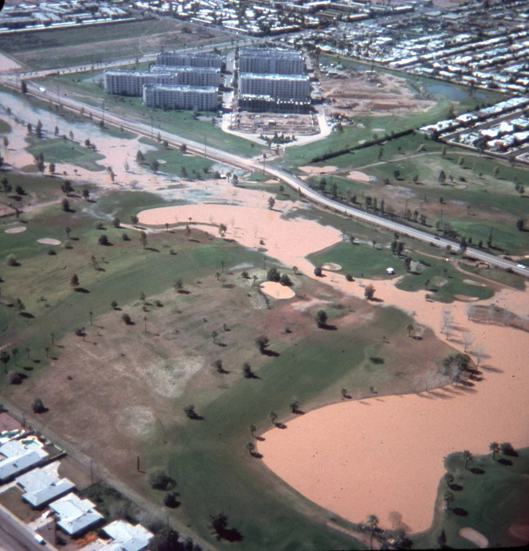 Aerial view of a golf course with multiple sand traps, water hazards, and mature trees, adjacent to a large residential complex with multiple buildings.