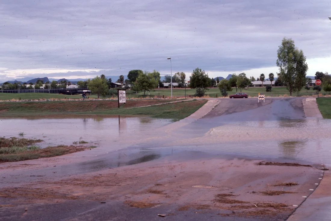 Flooded road and parking lot with water covering the ground, a stop sign visible, trees and a park in the background, and overcast sky.