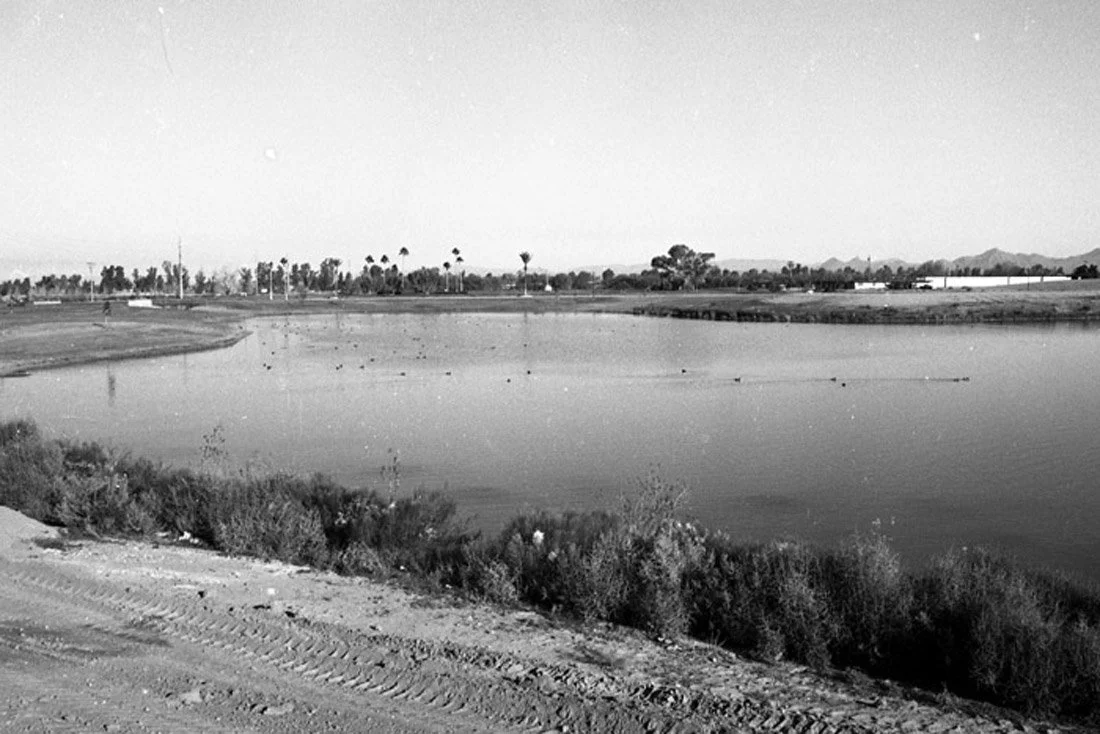 A black and white photo of a body of water, possibly a lake or pond, with a dirt path in the foreground and sparse vegetation around the water. In the background, there are trees, structures, and distant mountains under a clear sky.