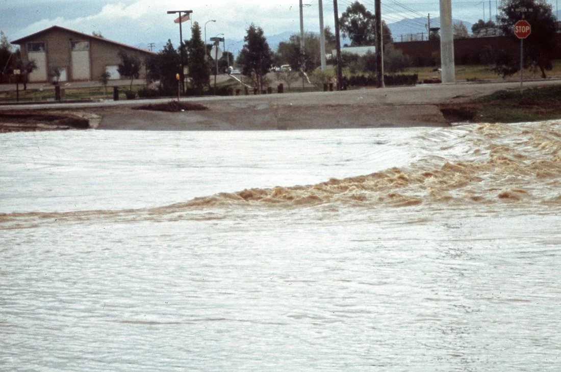 Flooded street with water covering the road, with a stop sign and buildings in the background.