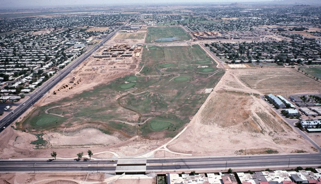 Aerial view of a golf course under construction with uneven terrain, patches of grass, and construction materials. Surrounding urban area with streets, buildings, and a highway in the foreground.