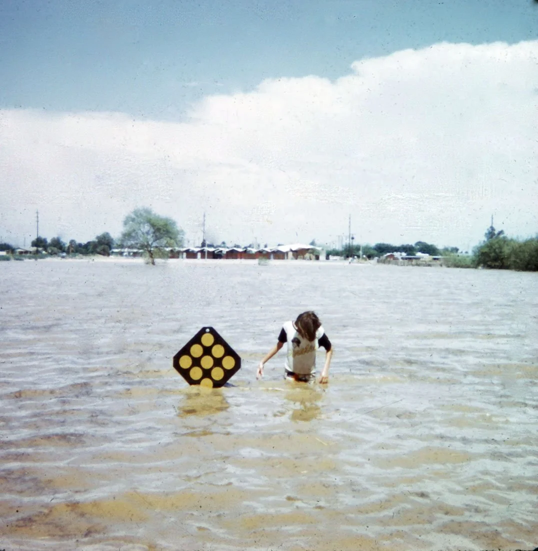 A person wades through floodwaters in a large open area with trees and houses in the background, holding a black and yellow object.