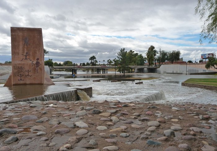 Flooded area with water flowing over rocks and a small waterfall, a monument with African art on it, and distant trees and buildings under a cloudy sky.