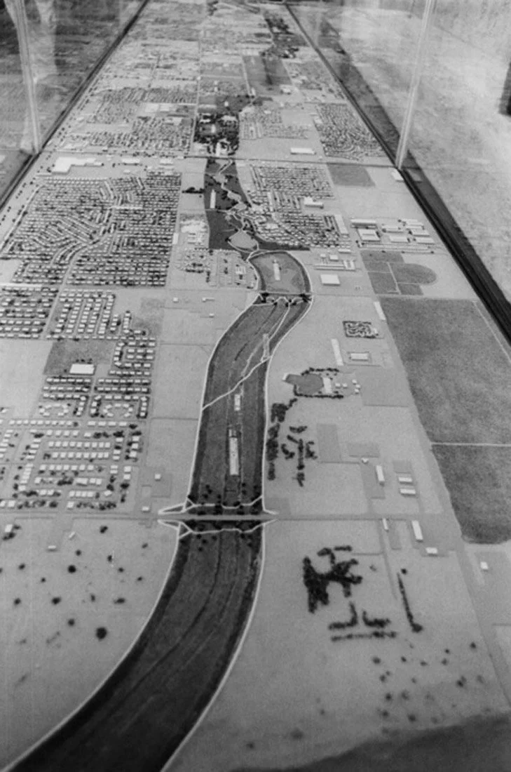 A detailed scale model or map of a city or town, displayed in a glass case, showing roads, waterways, residential, and commercial areas in grayscale.