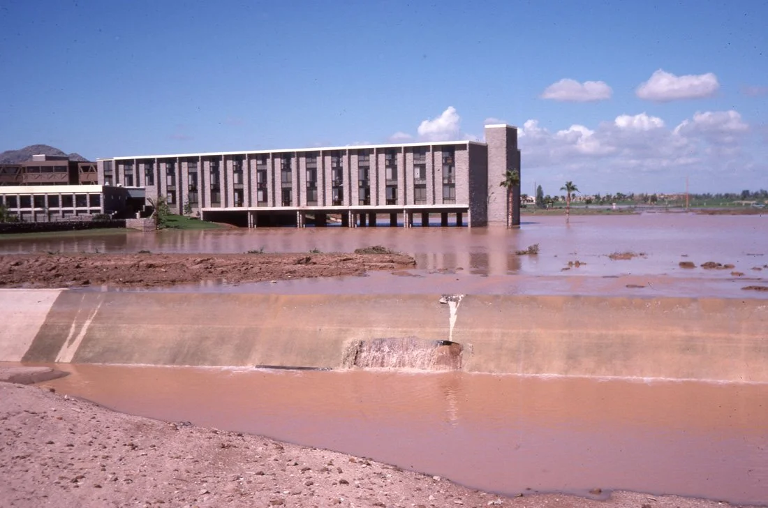A large, modern building sits on the edge of a flooded area with muddy water. The sky is blue with some clouds. The floodwaters cover the ground and reach near the base of the building.