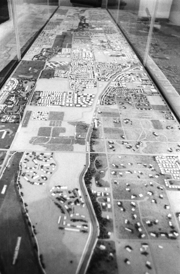 Black and white image of a glass display case containing a detailed miniature model of a suburban area with houses, roads, and green spaces.