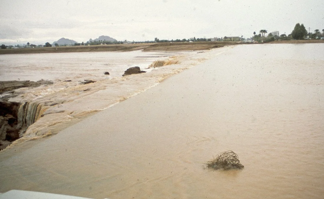 Flooded road with muddy water, damaged surface, and debris, in a rural area with trees and mountains in the background.