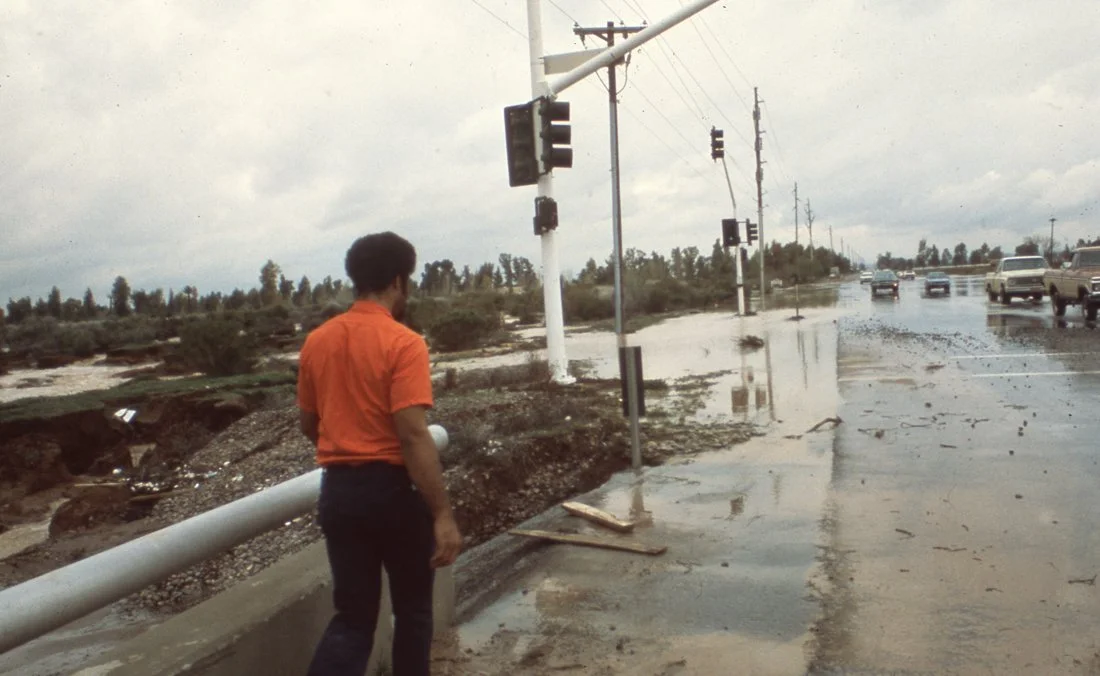A man in an orange shirt standing next to a fallen utility pole on a flooded street with damaged traffic signals and debris, with cars driving through the flooded area.