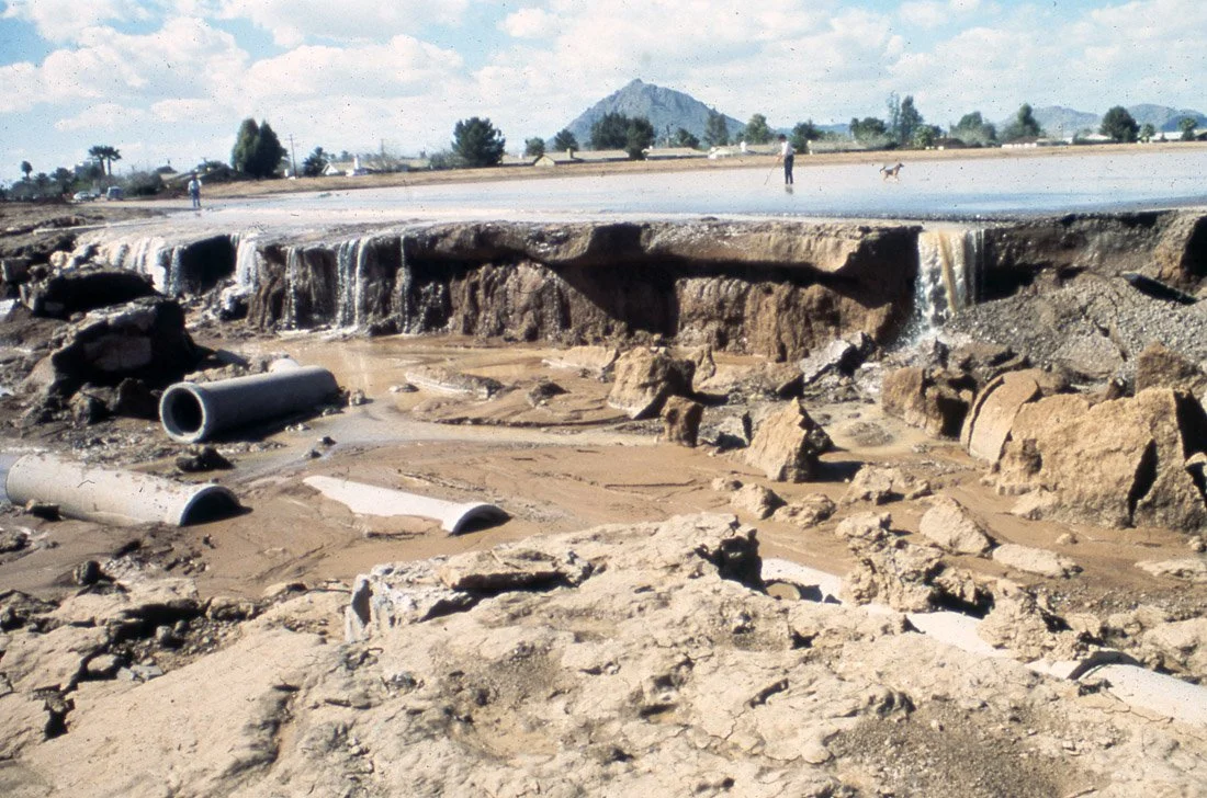 Flooded area with broken ground, mud, rocks, and scattered pipes, with a damaged road and distant hills in the background.