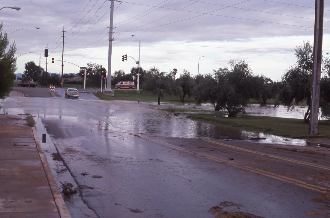 Flooded street with water covering part of the road, trees and utility poles in the background, overcast sky, and cars waiting at a traffic light.