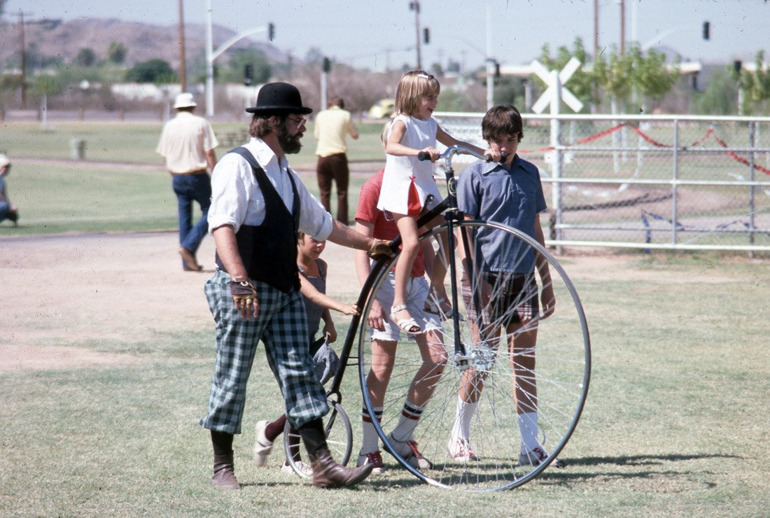 A young girl riding a penny-farthing bicycle with a person assisting her at a park on a sunny day.