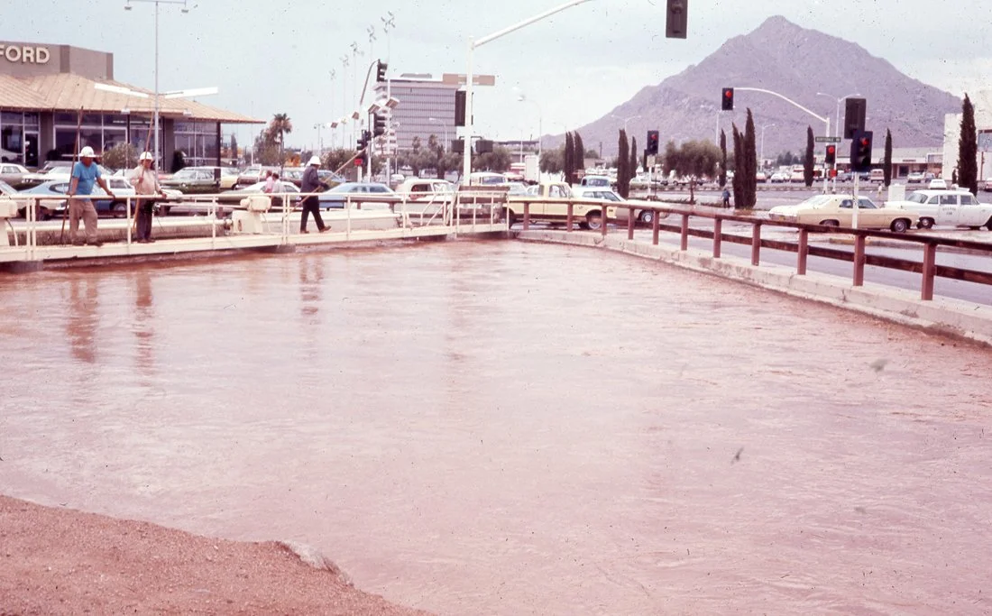 Flooded parking lot with cars and people walking on the sidewalk, traffic lights, mountains in the background.