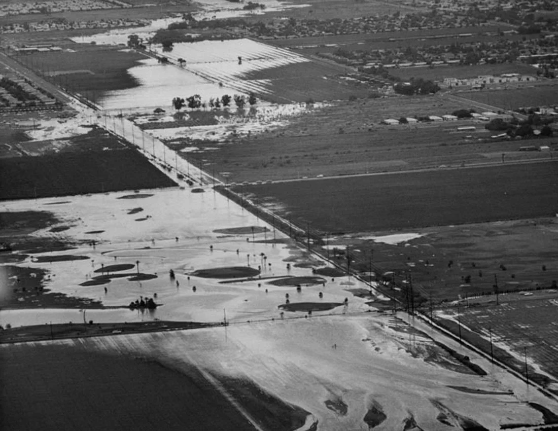 Aerial view of flooded fields and waterlogged ground, with several people walking across the area, along with some cars and trees in the distance.