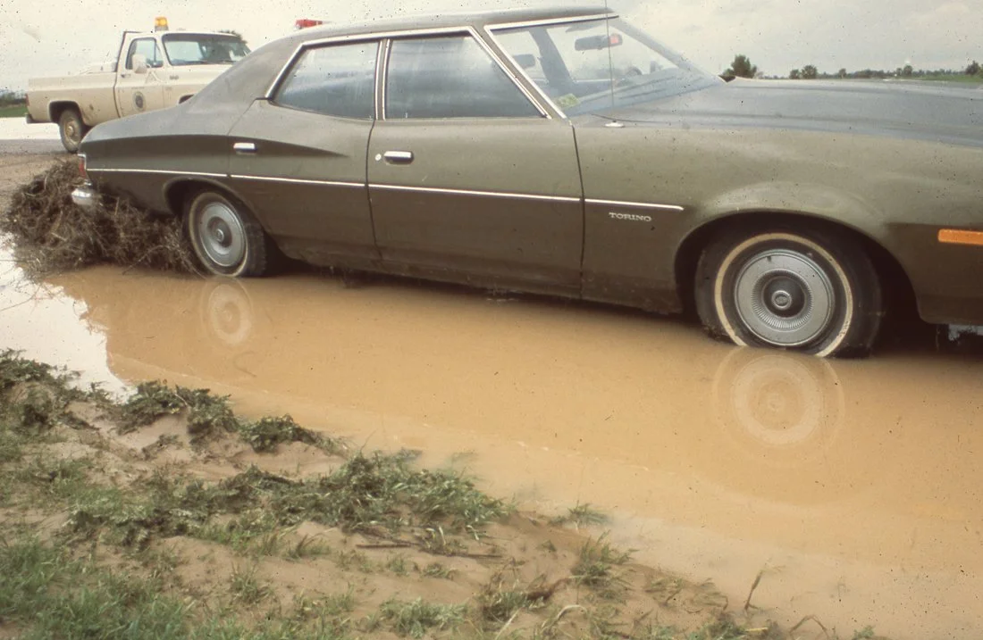 A vintage brown car partially submerged in water, with muddy puddles and grass nearby, and a beige utility vehicle in the background.