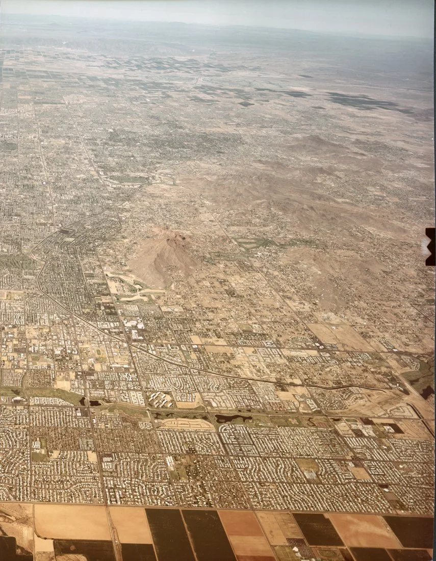 Aerial view of a city with a mix of urban and desert areas, showing streets, neighborhoods, and patches of farmland, with mountains or desert terrain in the distance.