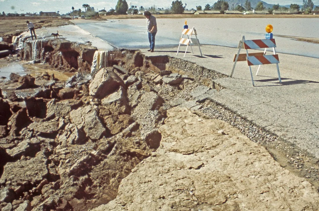A damaged road with a collapsed section, construction barriers, and a person walking along the edge with a dog in the background.