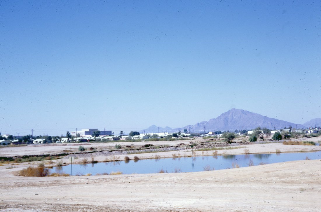 A desert landscape with a small body of water, distant buildings, and a mountain range in the background under a clear blue sky.