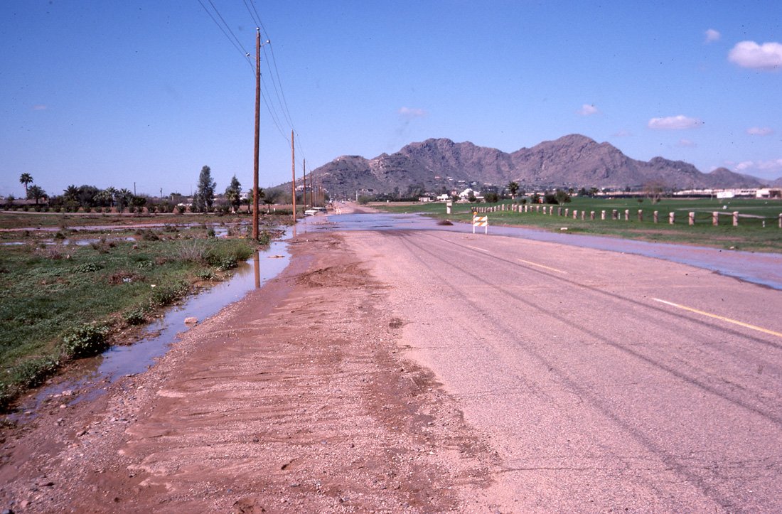 A rural road with puddles and muddy areas, utility poles on the side, green fields, and mountains in the background under a blue sky with scattered clouds.